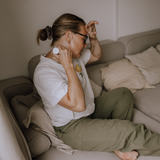 En kvinna sitter i soffan och applicerar en roll-on i nacken medan hon håller sig för pannan — som lindring mot spänningar eller huvudvärk.